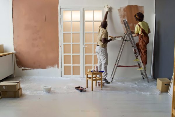 Couple painting interior walls while using ladder, brushes, and paint. Floor is covered with protective sheet, and tools and paint cans are scattered around